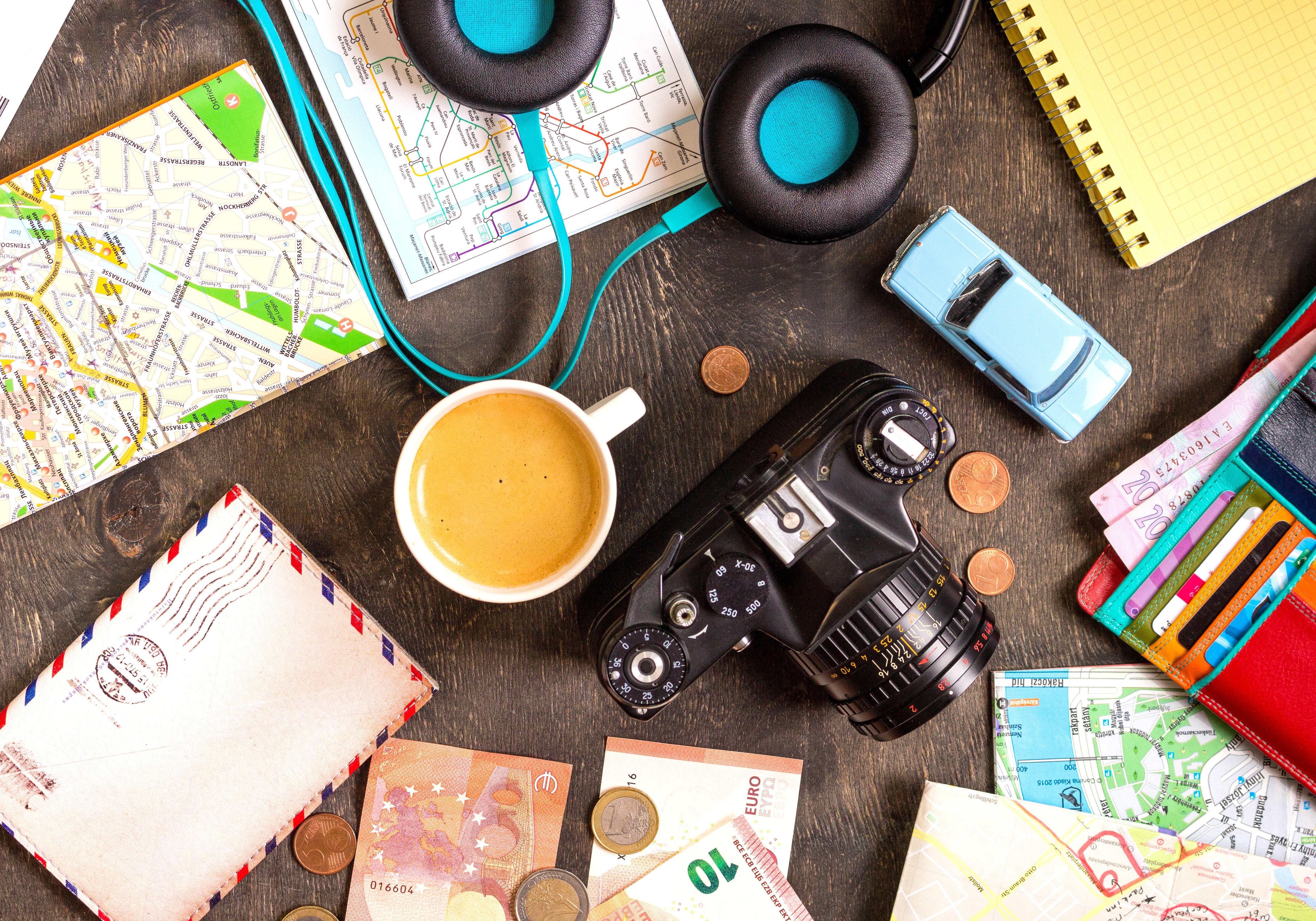 A collection of travel essentials on a black desk - camera, touristic maps, passport, toy car, coffee, headphones, wallet with credit cards, euro banknotes, and coins.