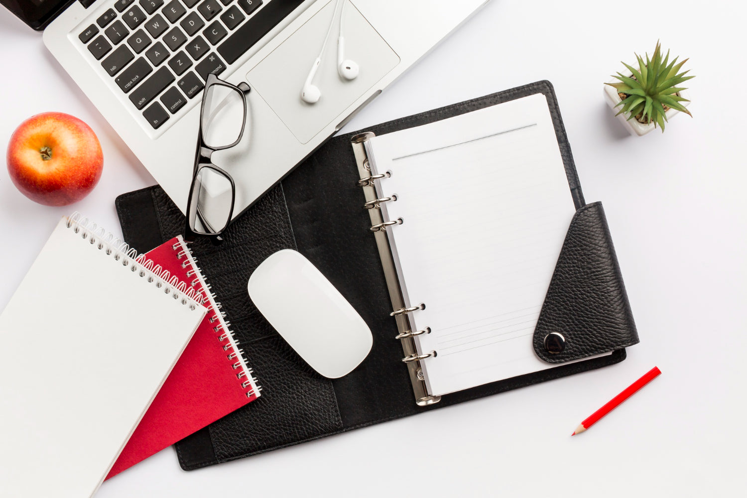 A composition of items on a white desk - red apple, diary, mouse, eyeglasses, earphones, pencil, and laptop.